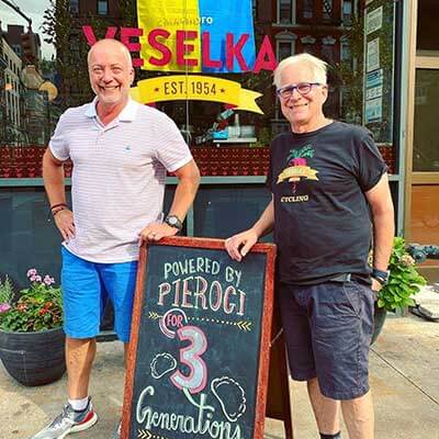 Jason and tom posing outside of Veselka 2nd ave with a "powered by pierogi for 3 generations" sandwich board in the summer 