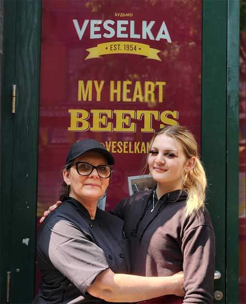 Olesia with her daughter in front of Veselka graphics on a side door, hugging. 