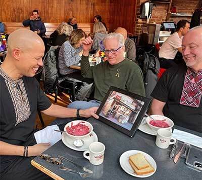 Jason and tom with bowls of borscht sharing an original photo of the Veselka location in second ave to a news reporter covering the work Veselka is doing to help Ukraine during the war